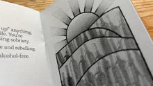 Load image into Gallery viewer, A close-up of an open book or booklet showing printed text on one page and a black-and-white sunburst illustration on the other, placed on a wooden surface.
