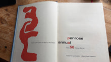 Load image into Gallery viewer, An open book lying on a wooden surface, showing a white page with a large abstract red graphic across the top and vertical red text reading 'annual penrose' on the lower page.
